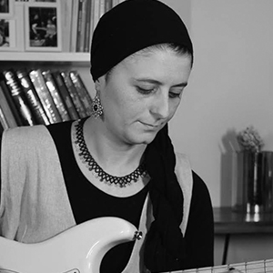 Rümeysa Çamdereli, beyaz tenli, 40'larında bir kadın. Uç kısmından siyah saçlarının bir bölümünün göründüğü beline kadar uzanan bir başörtüsü takmış. Bir kitaplık önünde oturarak gitar çalıyor.